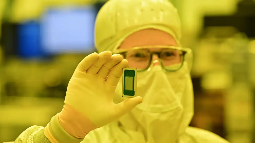 Person in a clean room suit holds a small computer chip up in gloved hand