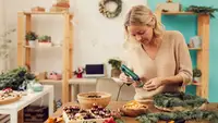 Woman using a hot glue gun to attach dried fruit to an evergreen Christmas wreath