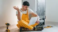 A man sits on the ground near a fridge, near a drill and some manuals. He's reading the fridge's warranty and putting up one hand in confusion at what he's reading.