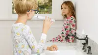 A girl and a boy wearing pajamas in a bathroom brushing their teeth.