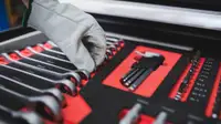 A work-gloved hand reaches into an open toolkit to grab a ratchet. The ratchets are organized in order of size. The tool kit is red with black detailing.