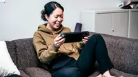 Person seated on couch holding portable gaming device and smiling at screen.