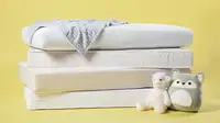 A stack of crib mattresses on a yellow background with two stuffed animals sitting on the floor next to them.