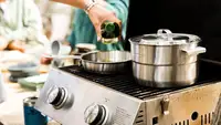 Stanley stainless-steel cookware is in use on a grill outdoors. Someone is pouring liquid from a bottle into a pan.