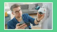 Person adjusting mounted smart security camera on wall indoors while holding smartphone.