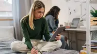 A female student sitting on a bed does homework in a dorm room while holding a tablet while another student sits at a desk behind her using a laptop.
