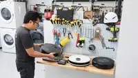 Person standing at work bench with assorted tools hanging along pegboard wall while inspecting the weight and dimensions of several small robot vacuums inside of testing lab with stacked washer and dryer combo.
