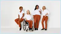 Four women, with one sitting in a wheelchair, are all wearing white T-shirts and Adaptive Denim Co. orange jeans.