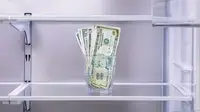 A glass with cash in it sitting on an empty refrigerator shelf.