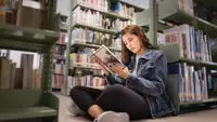 Student sitting on floor of the library, leaning against bookshelf and reading  a textbook