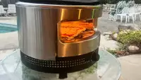 Solo Stove Pi Prime pizza oven with flames inside sitting on top of round table outdoors.