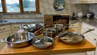 All Clad's D3 Stainless Steel Cookware set sitting on top of wooden island in the middle of modern kitchen.