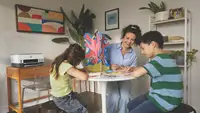 A woman and two children smile and work on a colorful arts and crafts project at a round table in a cozy, plant-filled home office with an HP printer in the background.