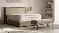 A modern bedroom featuring a white Saatva Classic mattress on a low-profile upholstered bed frame with warm wood accents, styled with neutral decor and soft natural lighting—an image highlighting the Saatva Memorial Day sale on luxury mattresses.