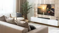 A bright, modern living room featuring an LG Dolby Atmos soundbar mounted beneath a large flat-screen TV on a light wood accent wall. The minimalist space includes a beige sectional sofa, white media console, indoor plant, and soft neutral decor, showcasing a clean and immersive home entertainment setup.