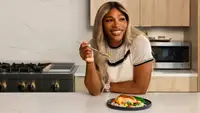 Serena Williams smiles while eating a Factor meal of blackened fish, grain salad, and green beans in a kitchen.
