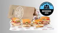 Three Jersey Mike's sub sandwiches loaded with deli meat, tomatoes, and shredded lettuce on a branded wrapper, with a Jersey Mike's paper bag in the background and a Readers' Choice Reviewed 2026 badge.
