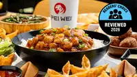 A close-up of a Pei Wei beef lo mein bowl with noodles, green beans, red peppers, and glazed beef on an orange branded background, with a Pei Wei cup visible and a Readers' Choice Reviewed 2026 badge.