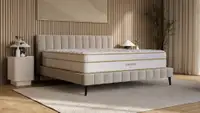 Saatva Classic innerspring mattress with Euro pillow top and gold trim on a beige upholstered bed frame in a warm wood-paneled bedroom.
