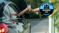 A barista handing an iced coffee drink through a drive-thru window to a customer in a car.