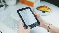 Hands holding an Amazon Kindle above a white table with an open book and pastry