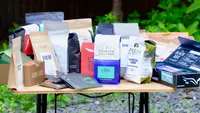 Over a dozen different bags of coffee beans arranged on a wood table outdoors.