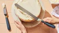 One hand holds a cake stand steady while the other spreads vanilla frosting across the cake using a large offset spatula.