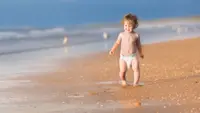 A baby in a diaper walking in the sand.