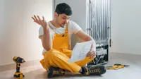 A man sits on the ground near a fridge, near a drill and some manuals. He's reading the fridge's warranty and putting up one hand in confusion at what he's reading.