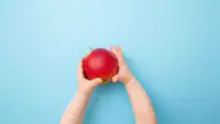 A toddlers hands extended holding a red apple against a light blue background.