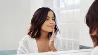 A person looks into a mirror while smiling and touching their face.