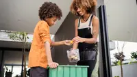 A brother and sister of color add food scraps to a large green compost bin