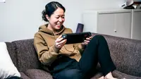 Person seated on couch holding portable gaming device and smiling at screen.