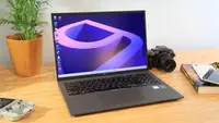 A laptop sits on a desk surrounded by a house plant, video game device, camera, and book.