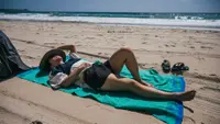 A woman reads a book while laying on a beach towel on the sand.