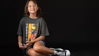 A tween, wearing a Naruto T-shirt and blue denim shorts from Stitch Fix's The Commons, sits on the floor with a black background.