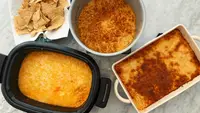 A bowl of tortilla chips and three pans of buffalo chicken dip laid out on a marble counter.