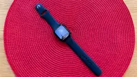 The Apple Watch SE with black band sits on a circular red background on a table.