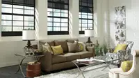View of a lovely studio apartment living space: a glass coffee table, abstract art on the wall, white walls, black smart blinds, a white decorative lamp, gold throw pillows, and a white rug.