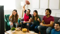 Five people sitting on a couch in the living room watching football on a flat screen TV