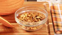 A clear bowl filled with pumpkin seeds on a wooden cutting board.