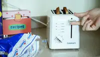 A Cuisinart 2-slice toaster is being shown in close-up. A hand demonstrates the toaster's operation; two slices of toast are ready to be retrieved from the slots.