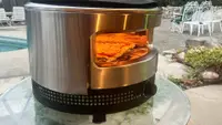 Solo Stove Pi Prime pizza oven with flames inside sitting on top of round table outdoors.