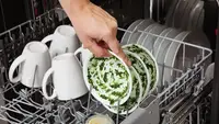 A person loading a bowl covered with spinach puree into a dishwasher that also has plates and mugs in it.
