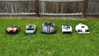 Five robot lawn mowers lined up together in the green grass with a fence behind