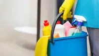 A person holds the handle of a bucket filled with bathroom cleaning supplies.
