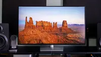 A widescreen monitor displays a high-resolution image of red rock formations in a desert landscape, framed by black studio speakers on a sleek desk setup.