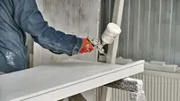 A person in protective clothing spraying white paint from a paint gun onto a piece of wood