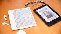 Two e-readers sitting on a wooden surface with a pair of glasses