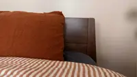 An orange pillow sits on the end of a bed near a wooden bedframe